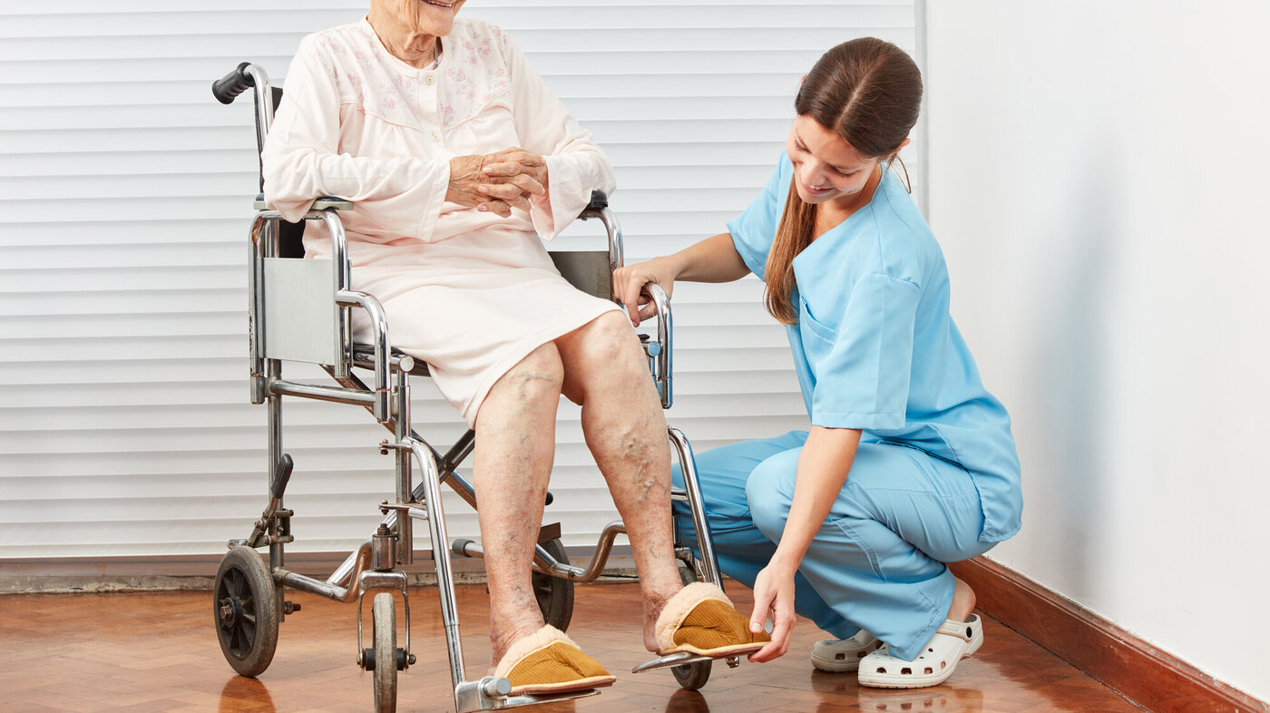 Bildet viser en sykepleier som sitter på golvet og hjelper en eldre dame i en rullestol.