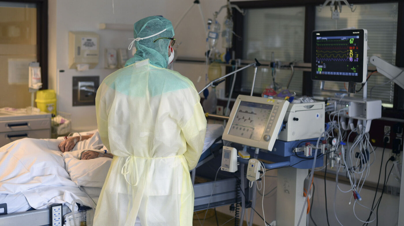 Bildet viser ryggen på et helsepersonell i smittevernutstyr, som står ved en pasientseng