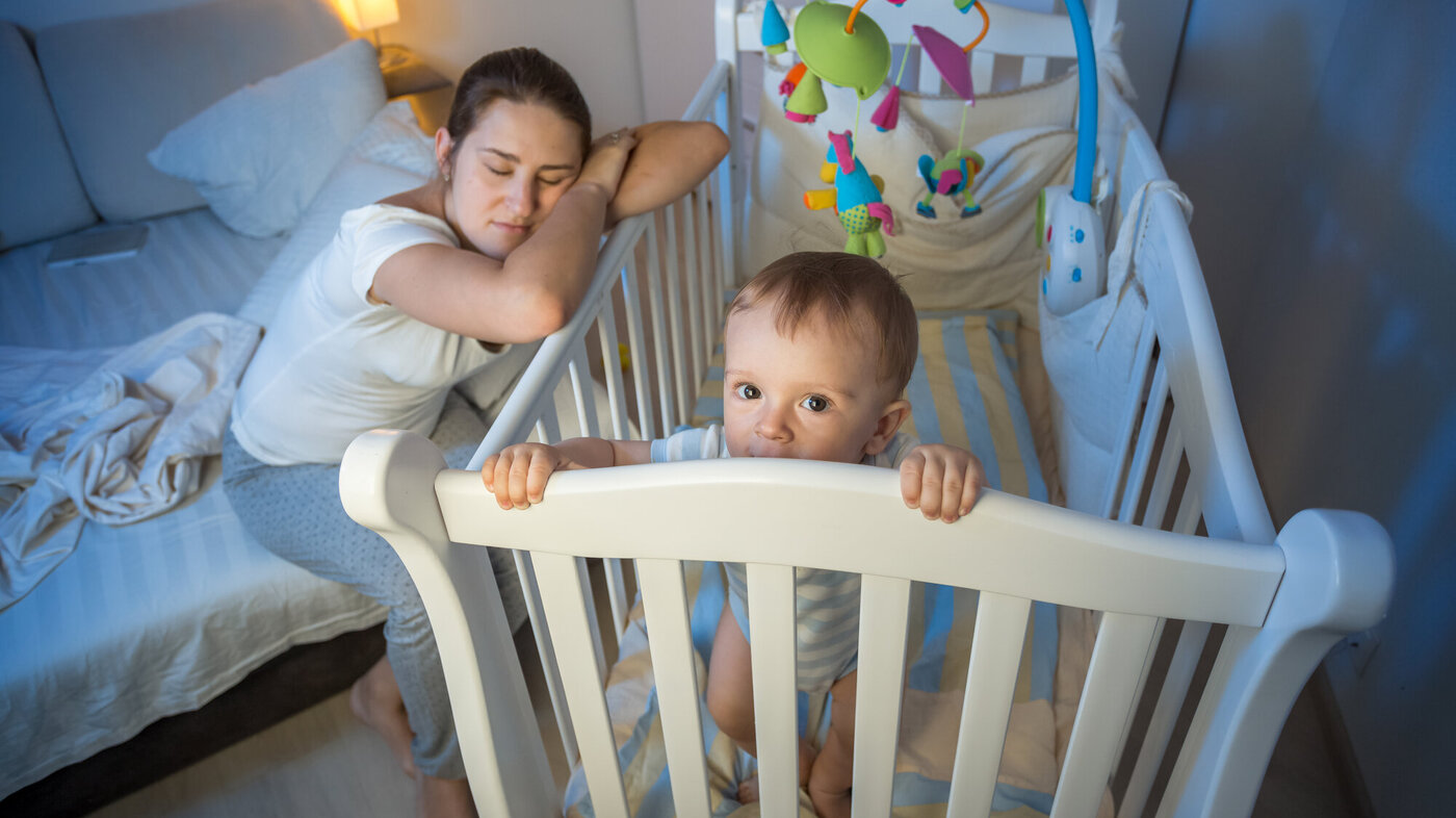 Bildet viser en mor som halvsover over barnesenga, mens barnet står i senga, lys våkent