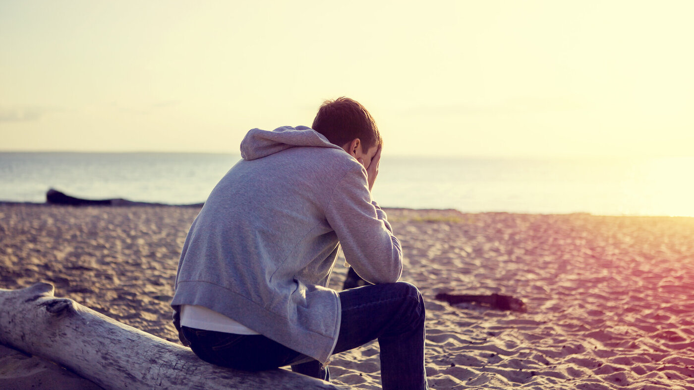 Bildet viser en ung mann som sitter på stranda med hodet i hendene