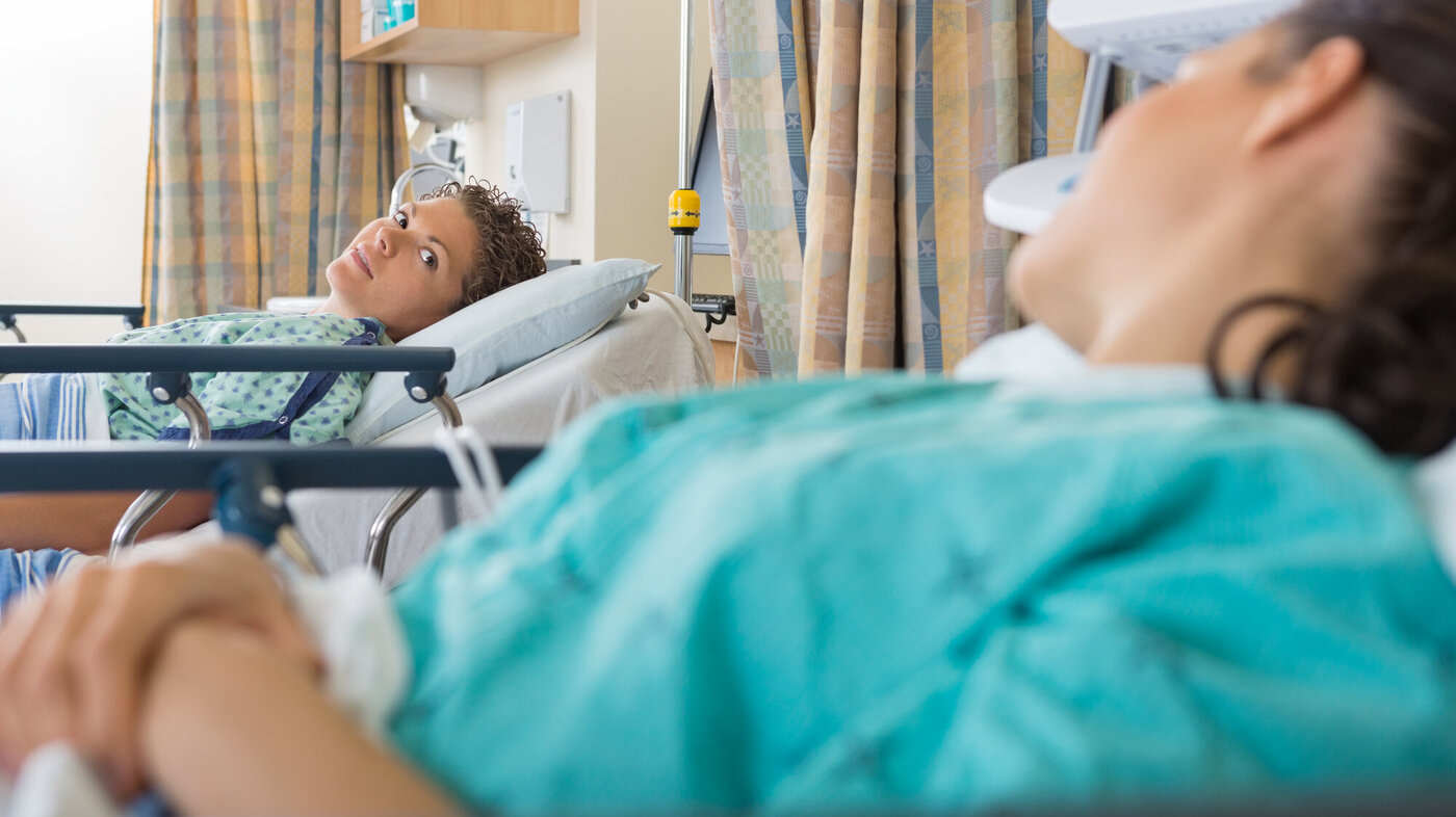 Bildet viser to kvinnelige pasienter som ligger i hver sin seng i operasjonsklær og ser på hverandre