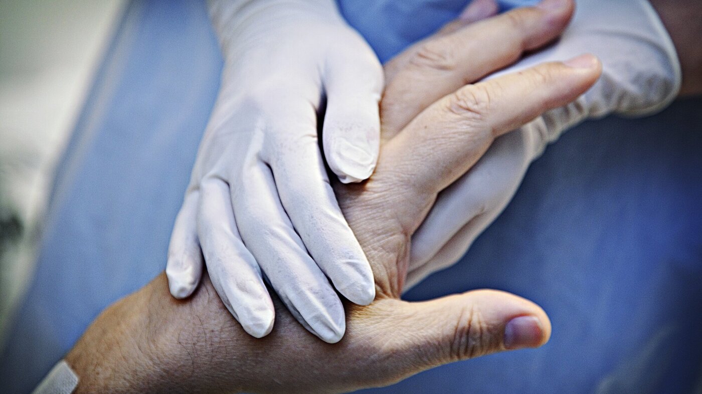 Bildet viser et par hender med plasthansker som holder hånda til en gammel person