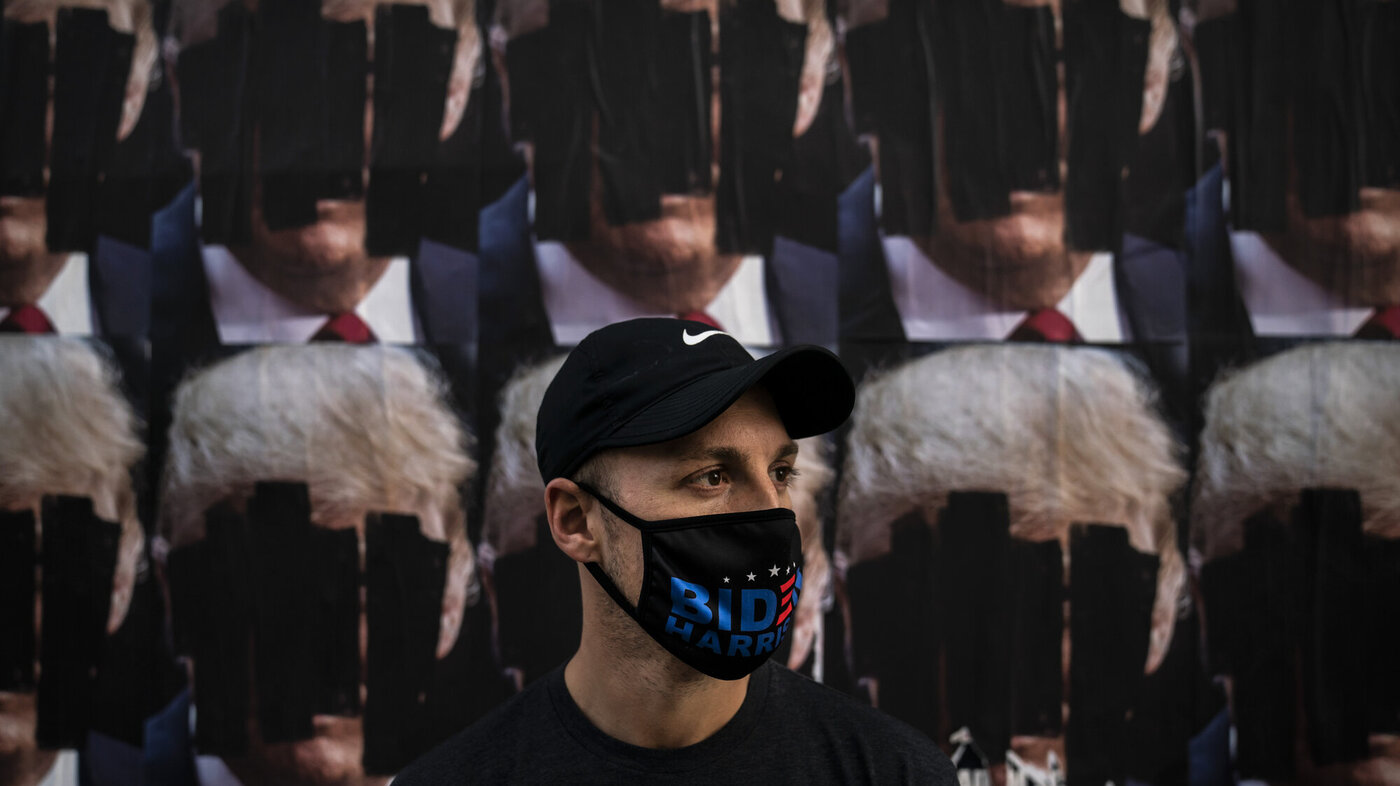 Bildet viser en mann med maske som det står Biden på, foran nedtusjete plakater av Trump.