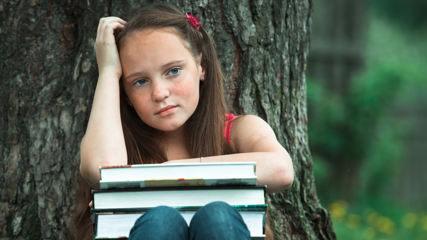 Bildet viser en ung jente som sitter ved et tre med en stabel bøker på knærne