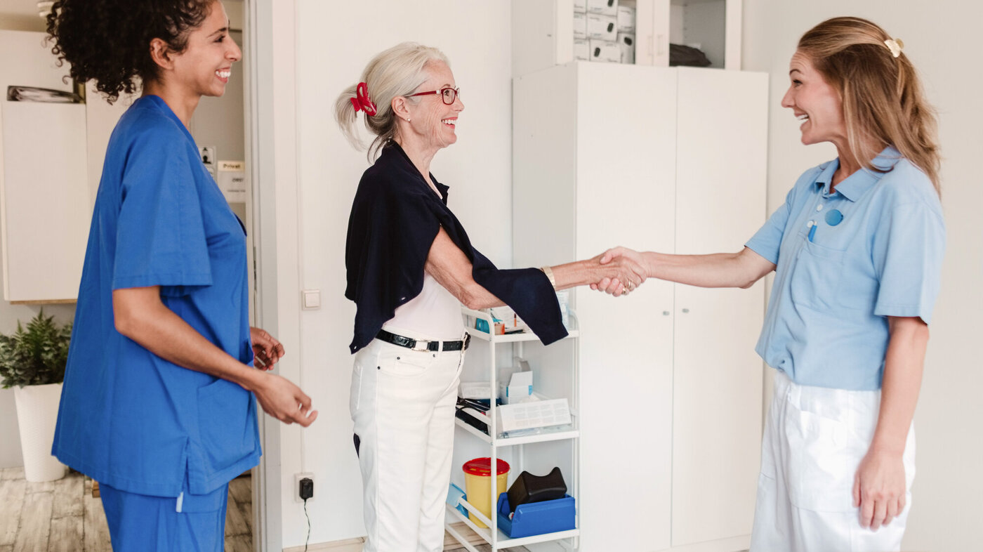 Bildet viser en eldre kvinne som tar et helsepersonell i hånden mens at annet helsepersonell ser på