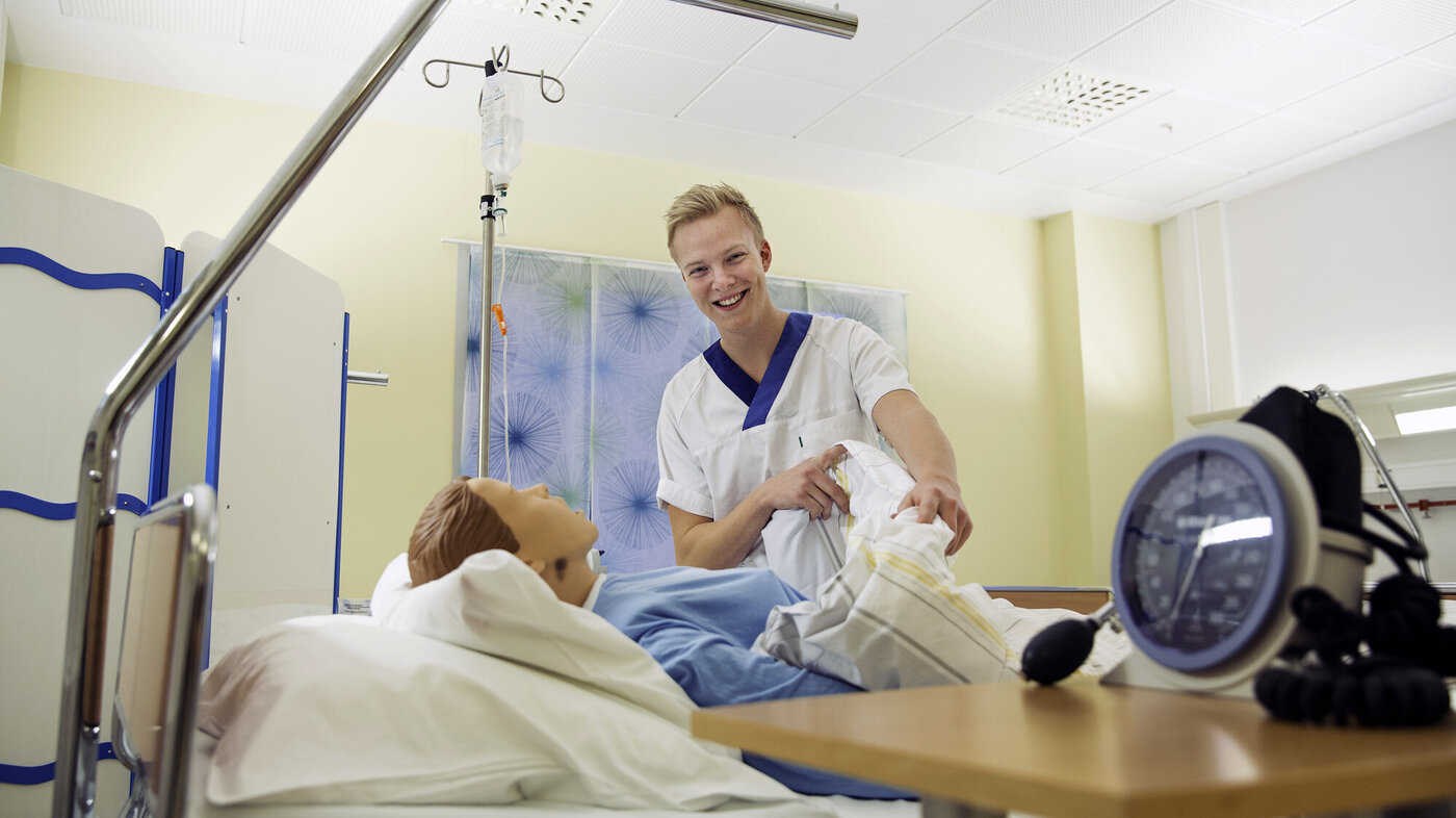 Bildet viser en sykepleierstudent som står ved siden av en seng hvor det ligger en simuleringsdukke