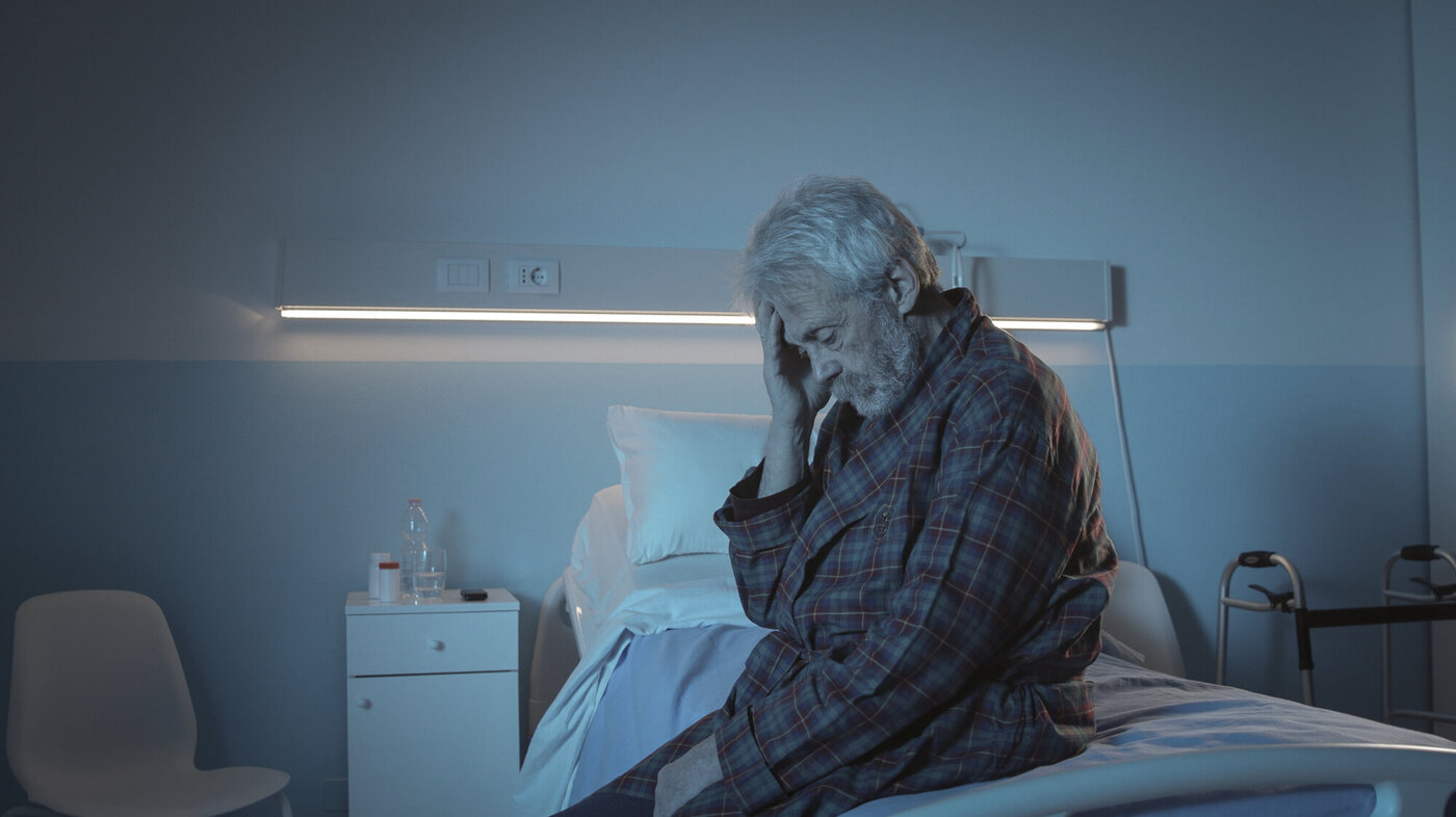Bildet viser en eldre mann som sitter på sengekanten med hodet i hendene