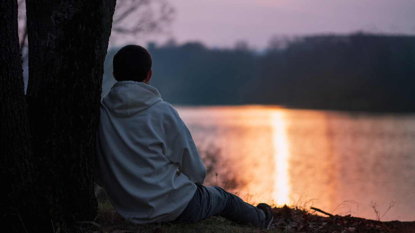 Bildet viser en ung mann sett bakfra som sitter ved et tre og ser ut på en solnedgang over et vann