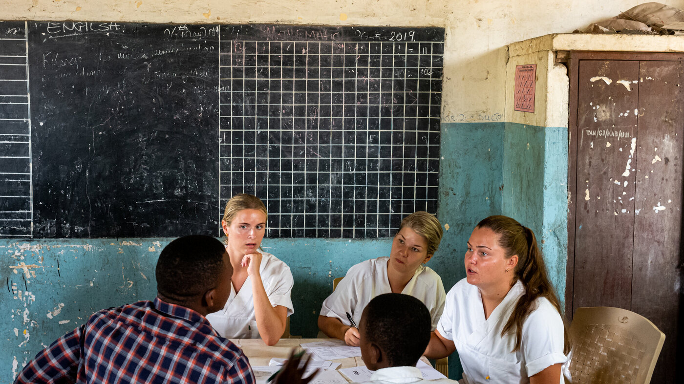 Bildet viser tre sykepleierstudenter fra Norge som er i Tanzania. De sitter og snakker med to afrikanske menn.