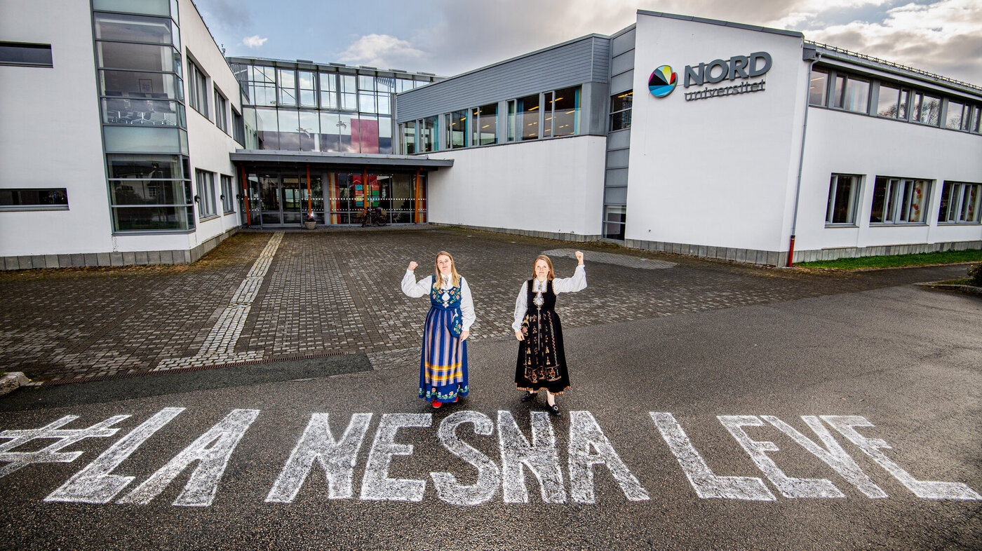 Bildet viser to bunadskledte mennesker utenfor høgskolen i Nesna, på bakken er det skrevet "La Nesna leve".