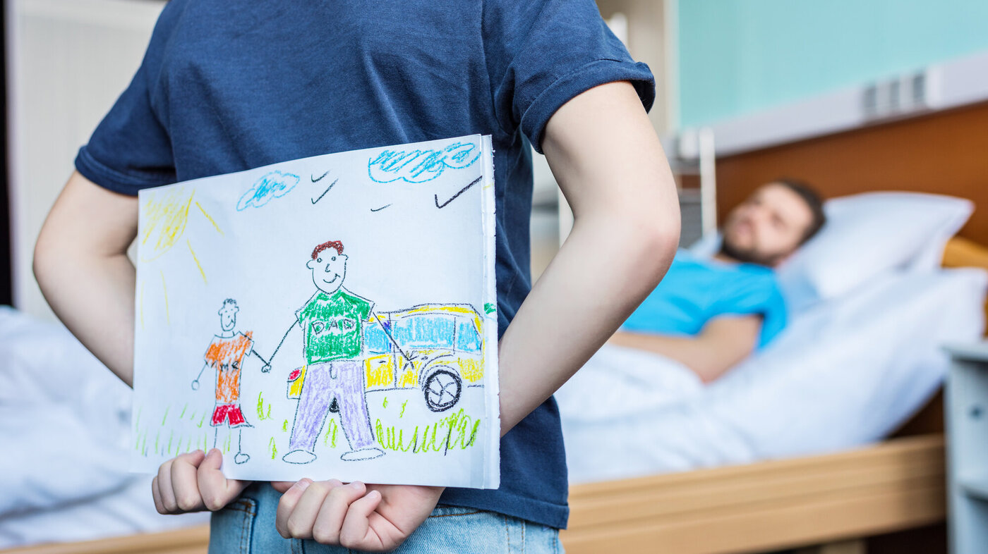 Barn gjemmer tegning bak ryggen ved fars sykeseng.