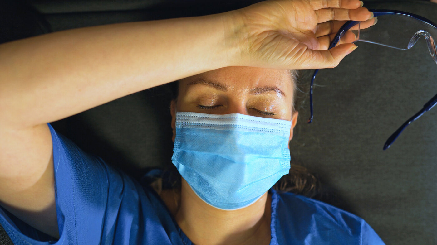 Bildet viser en trøtt sykepleier med munnbind som ligger på gulvet med armen over pannen
