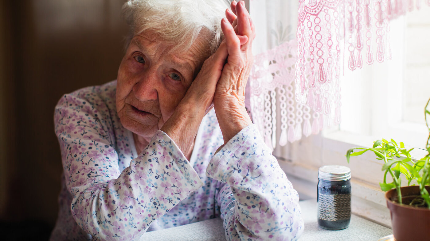 Bildet viser en trist, gammel dame som lener hodet mot hendene sine. Hun sitter ved kjøkkenbordet med et pilleglass foran seg.