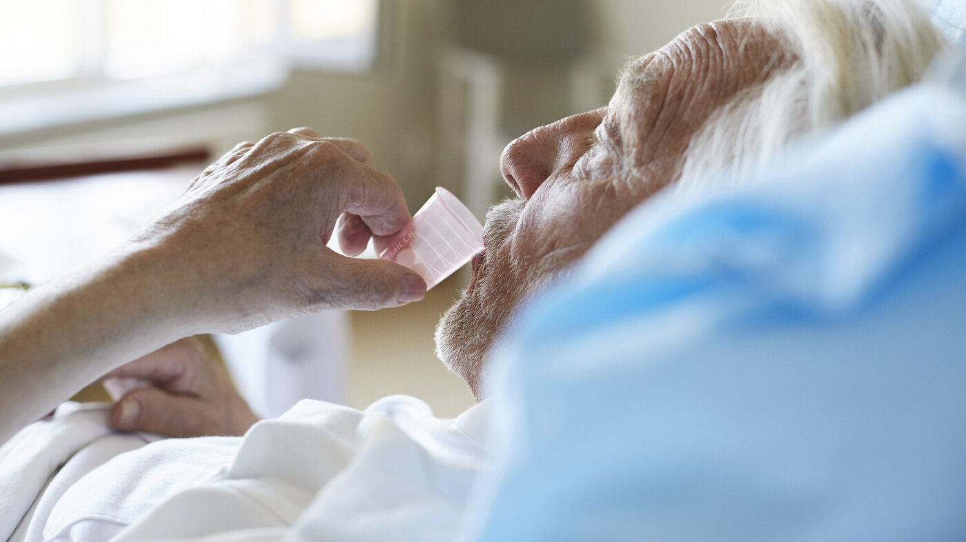 Bildet viser en eldre, syk mann i senga som drikker medisin fra et plastglass