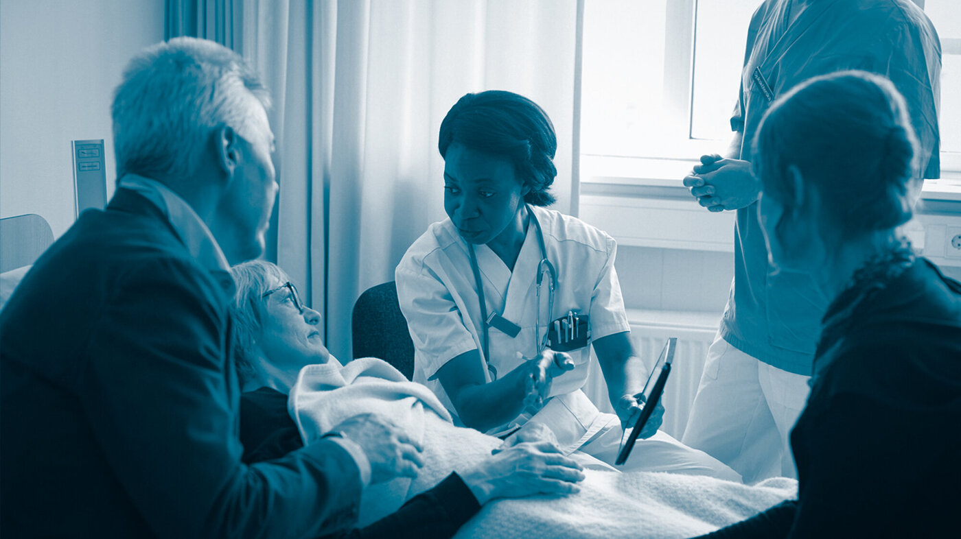 Bildet viser en sykepleier som forklarer noe til en pasient i senga og pasientens pårørende som står rundt