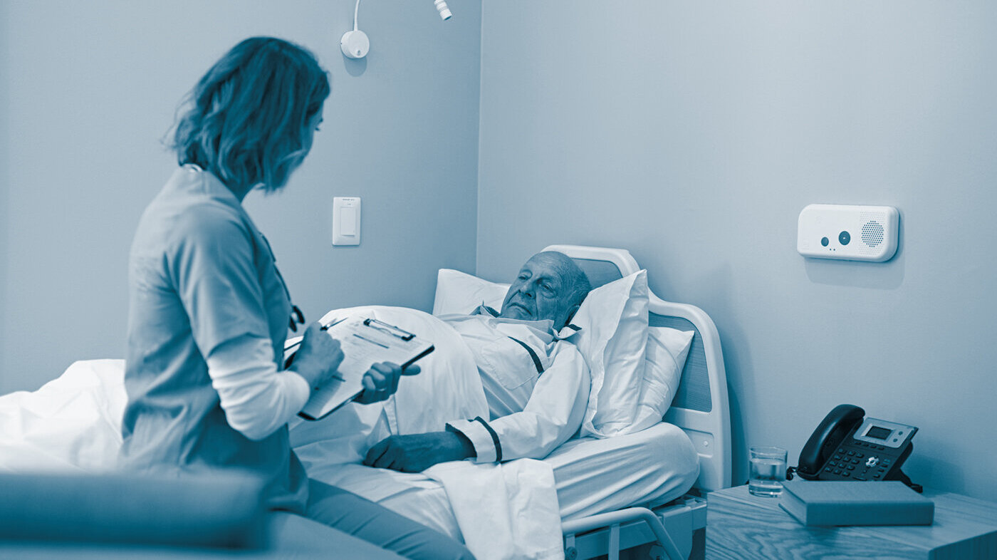 Bildet viser en sykepleier som sitter ved sengen til en eldre mann. Hun holder et skjema i hånda.