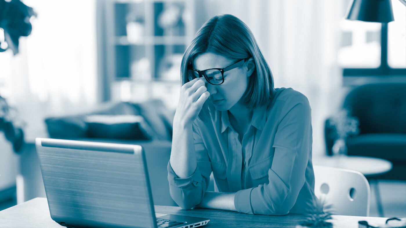 The photo shows a woman sitting at home with a laptop in front of her. She looks tired and is wiping her eyes.