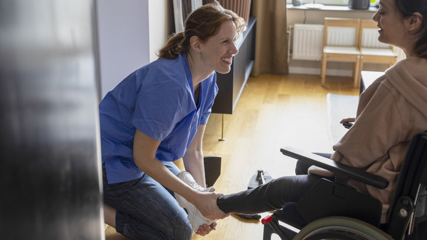 Bildet viser en kvinnelig sykepleier som hjelper en rullestolbruker på med skoene