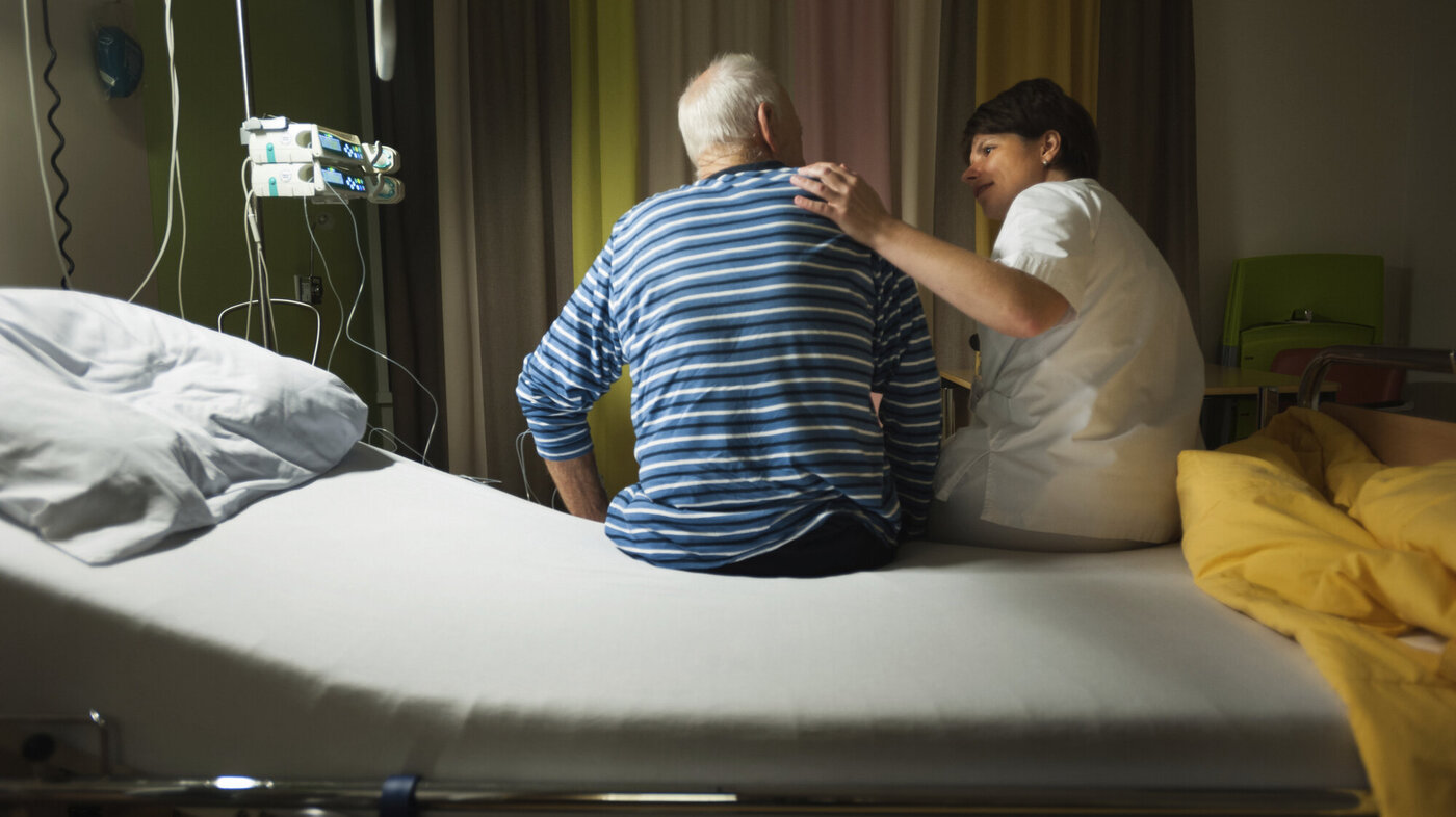 Bildet viser en sykepleier som sitter på sengekanten sammen med en eldre pasient