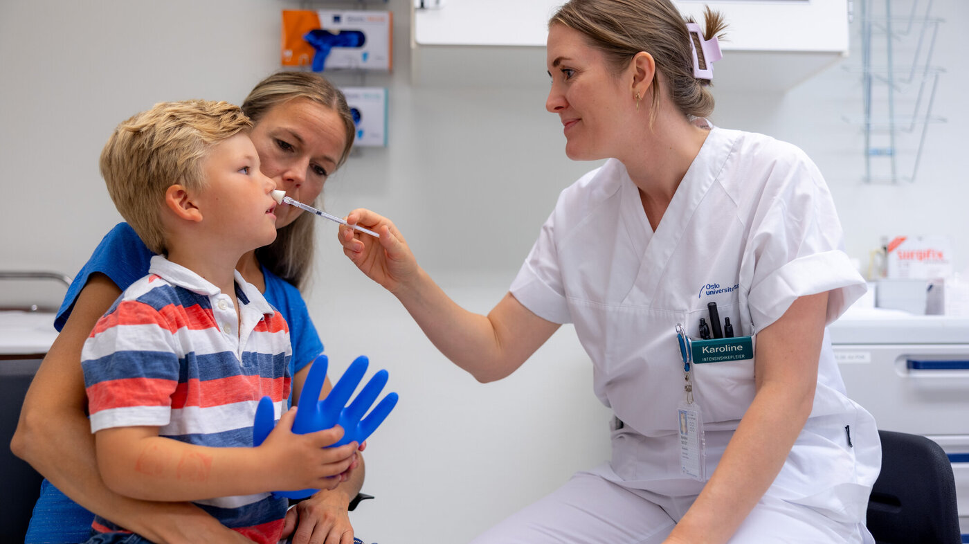 Bildet viser en liten gutt på fanget til sin mor. En sykepleier gir ham nesespray inn i det ene neseboret