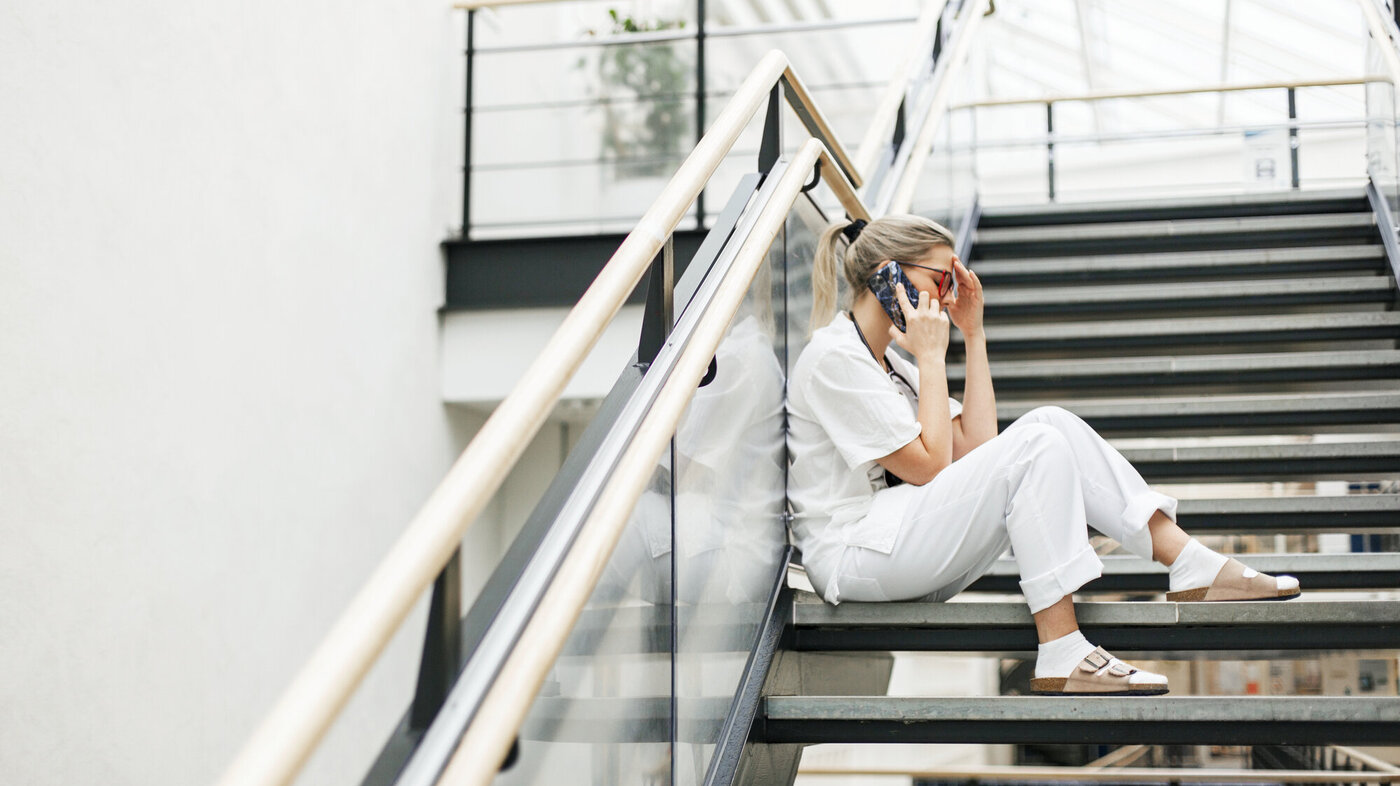 Bildet viser en sykepleier som sitter i en trapp, snakker i telefonen og ser sliten ut