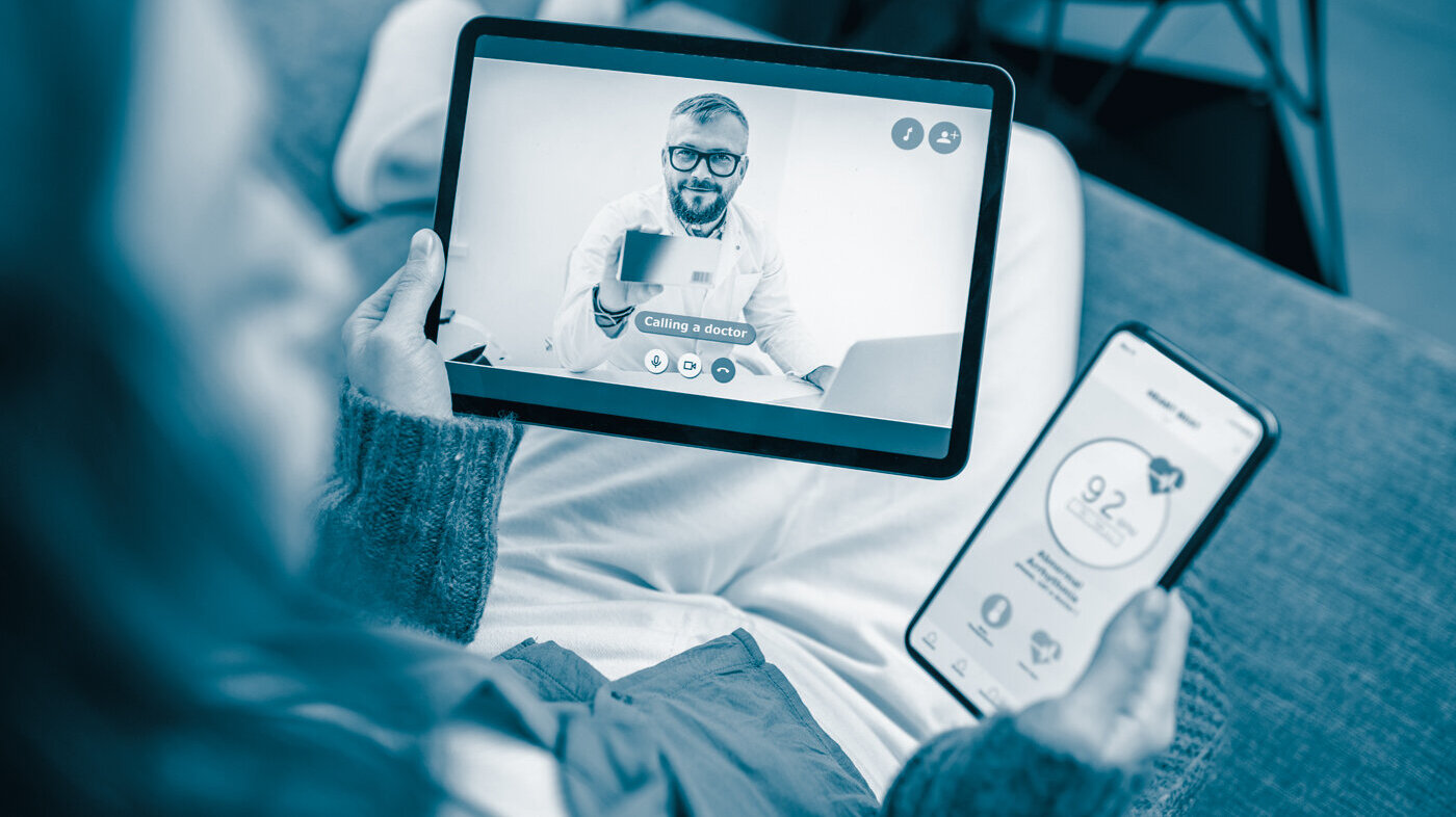 Digital home monitoring The photo shows a woman sitting in a sofa talking to a doctor via her Ipad. In the other hand she is holding a mobile phone where she is sending her vital data via an app