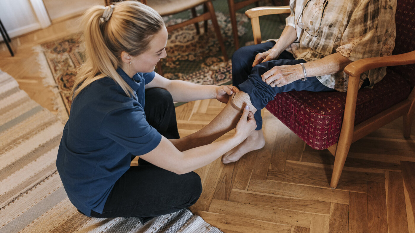 Hjemmesykepleie Bildet viser en hjemmesykepleier som skifter strømper på en eldre dame som sitter i stolen i hjemmet sitt.