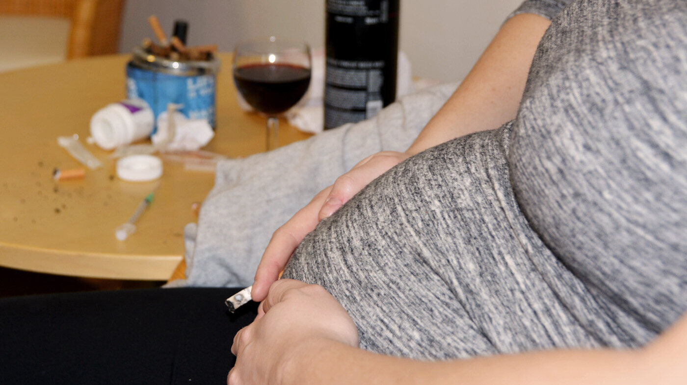 Rusmiddelbruk under graviditet Bildet viser magen til en gravid dame som sitter i en stol. I hånda holder hun en sigarett. På bordet bak står et askebeger fullt av sneiper og et glass med rødvin. Noen pilleglass ligger veltet på bordet.