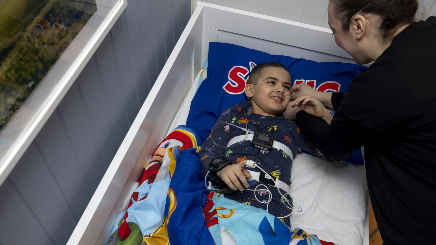 The photo shows a boy lying in his bed. Tools for sleep testing and monitoring are equipped on the boy. His mother is bending towards him, both are smiling