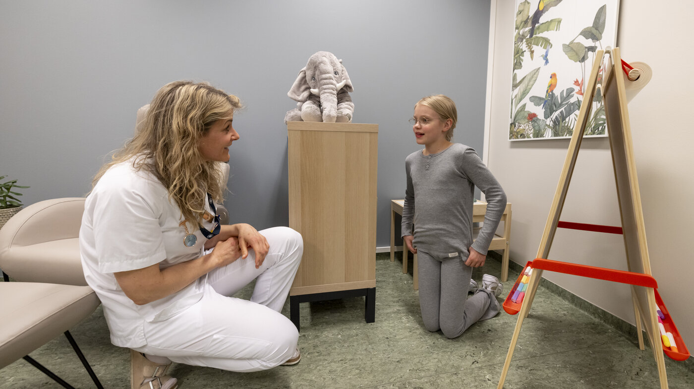 The photo shows a nurse squatting in front of a girl in a room at the hospital with a toy elephant and an easel.