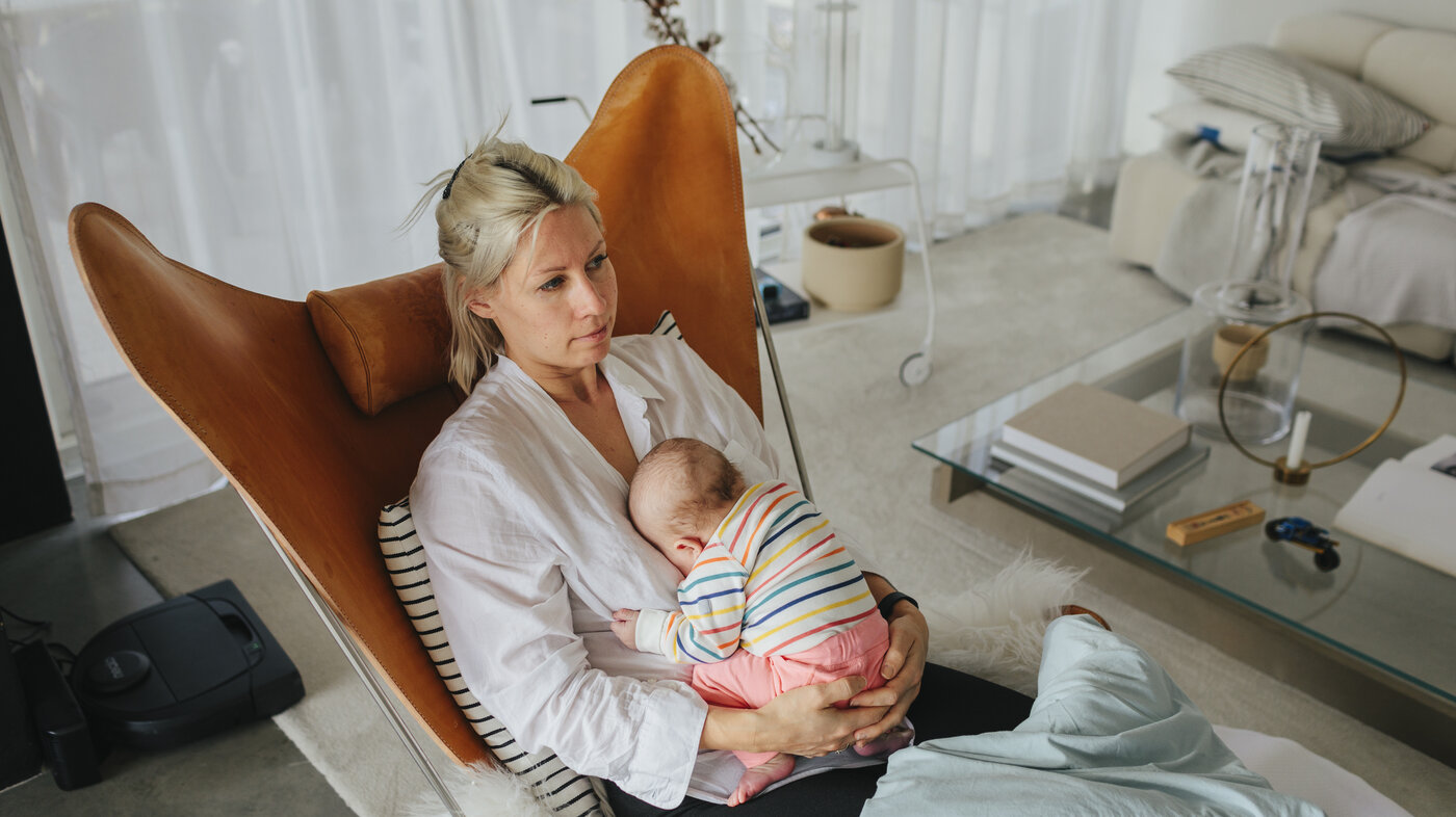 The photo shows a young mother sitting in a chair in her living room. She looks tired and holds her newborn baby in her lap.