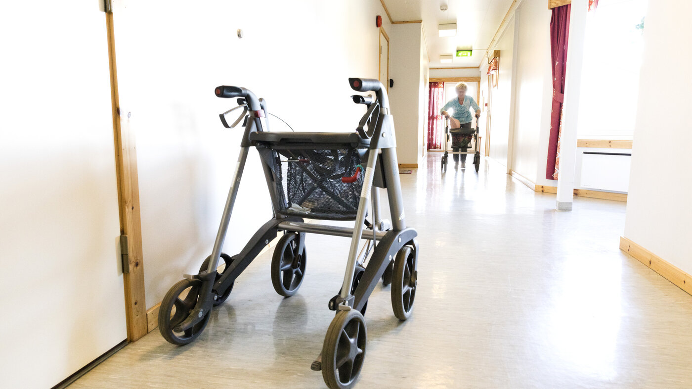 Bildet viser korridor med gåstol og en eldre dame som kommer gående - også med gåstol