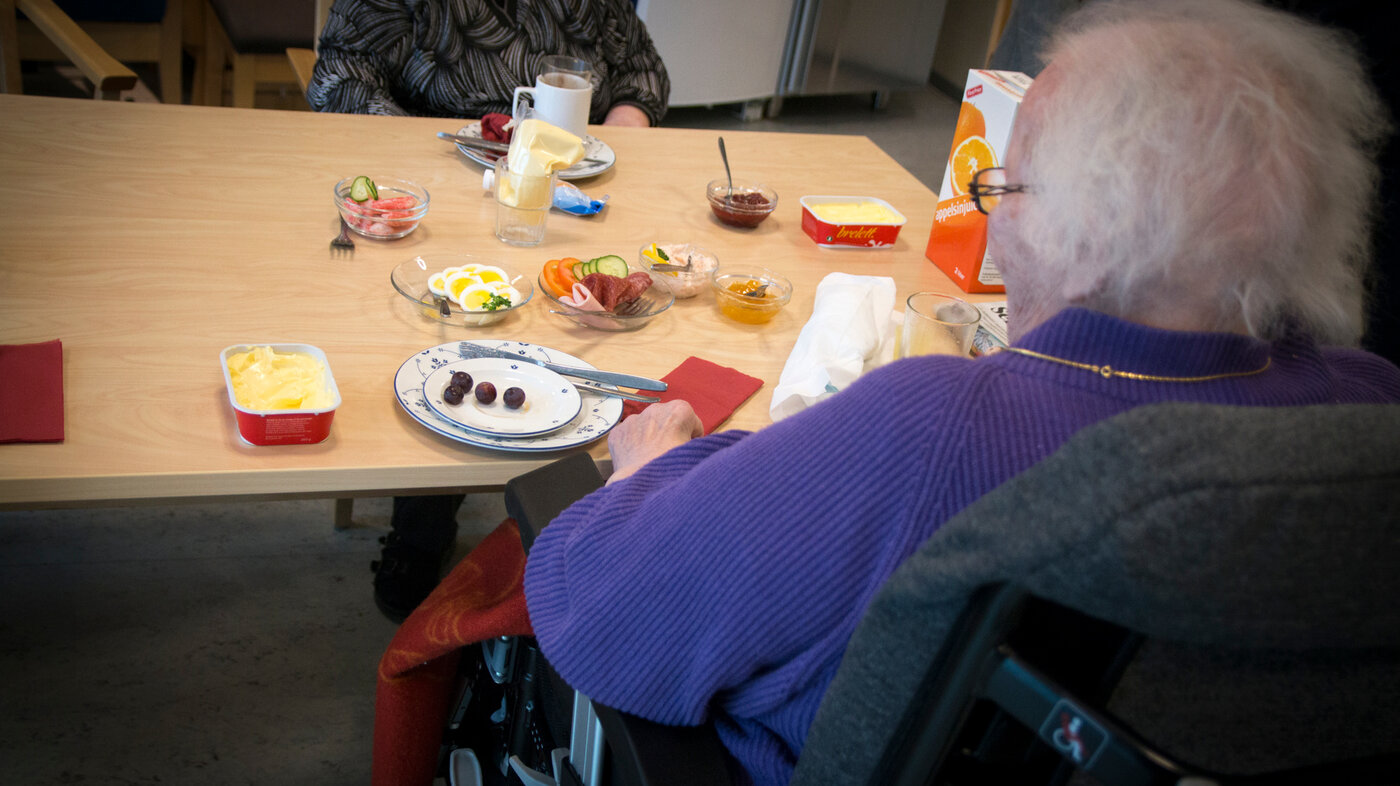 Bildet viser en eldre person som sitter og spiser mat med ryggen til