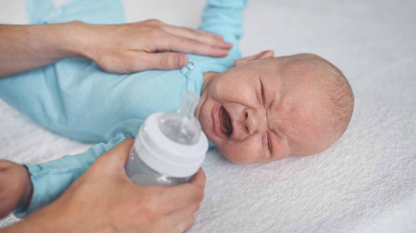 The photo shows a crying infant. A soothing hand lies comforting on the baby's stomach. The other hand is holding a bottle ready to feed the baby