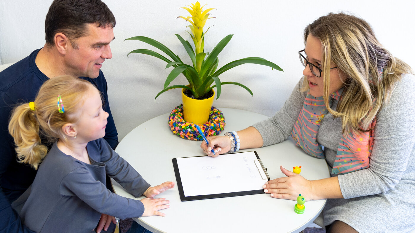 The photo shows a father with his daughter on his lap.  They are sitting at a table opposite a woman leaning towards them. She has a notebook in front of her.