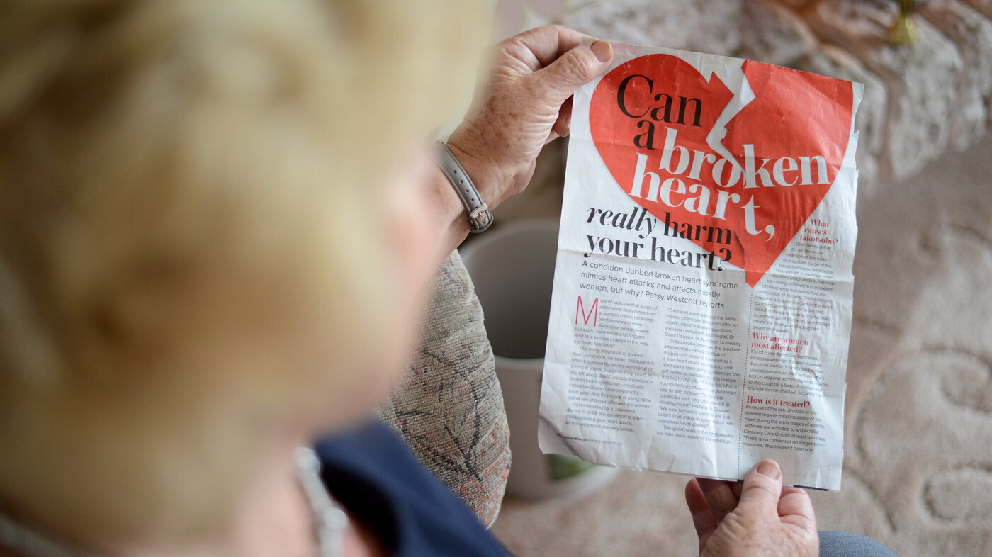 Bildet viser en eldre kvinne som holder opp et avisutklipp hvor det står "can a broken heart really harm your heart?"