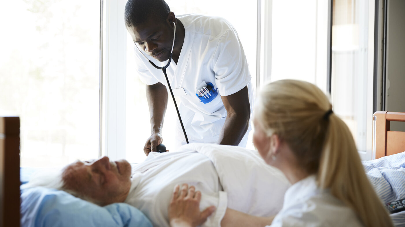 The photo shows an older patient lying in a hospital bed. A male nurse is bending towards him listening with a stethoscope. A female nurse is sitting beside the bed.