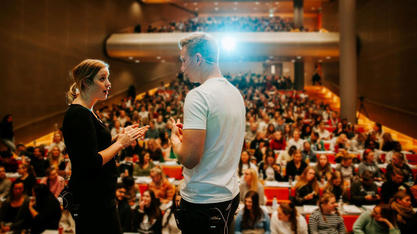 Bildet viser to studenter som står på scenen i et auditorium