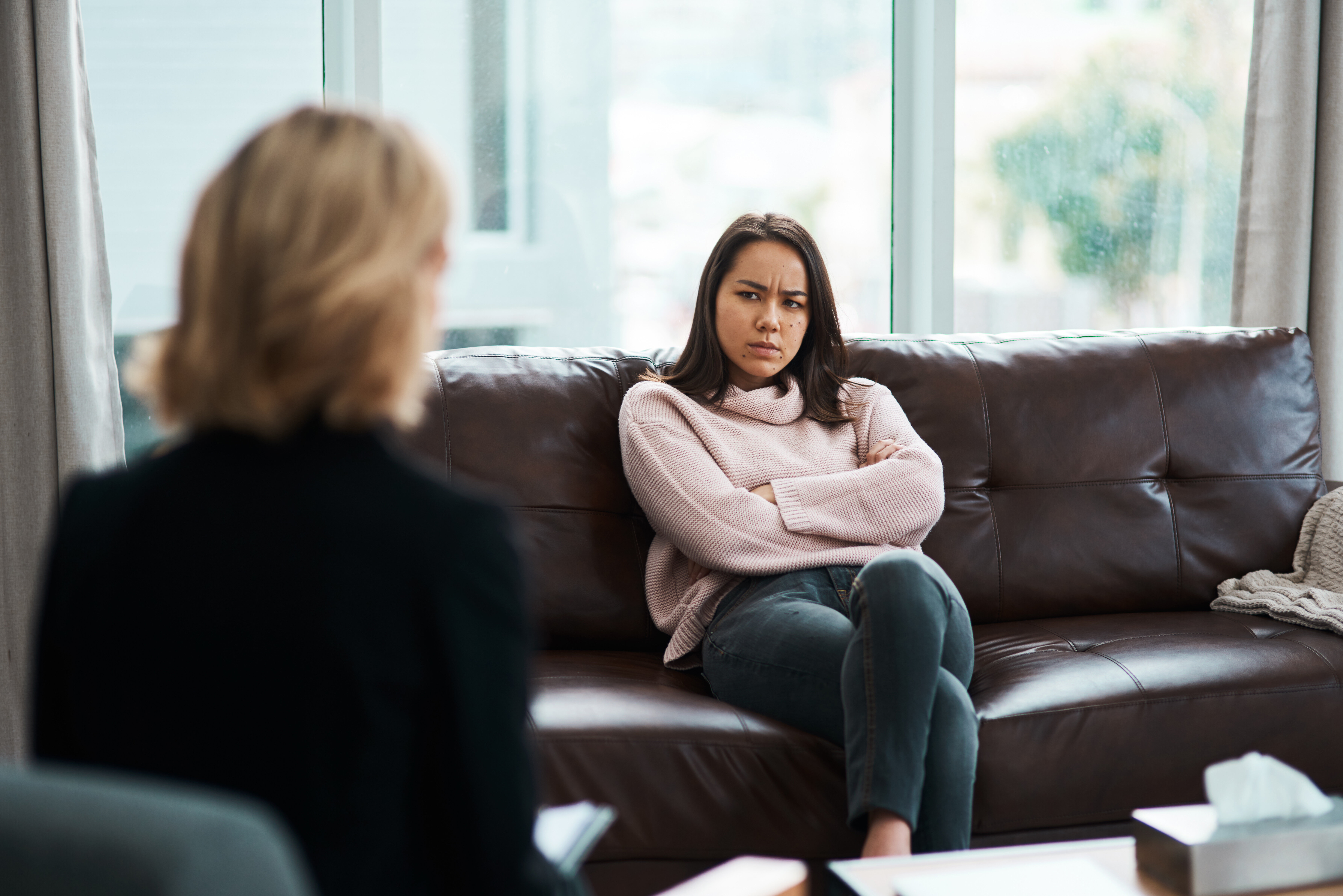 Bildet viser en person som sitter med ryggen til. Overfor henne sitter en ung jente som ser sint ut. Hun har armene i kors og et sint uttrykk