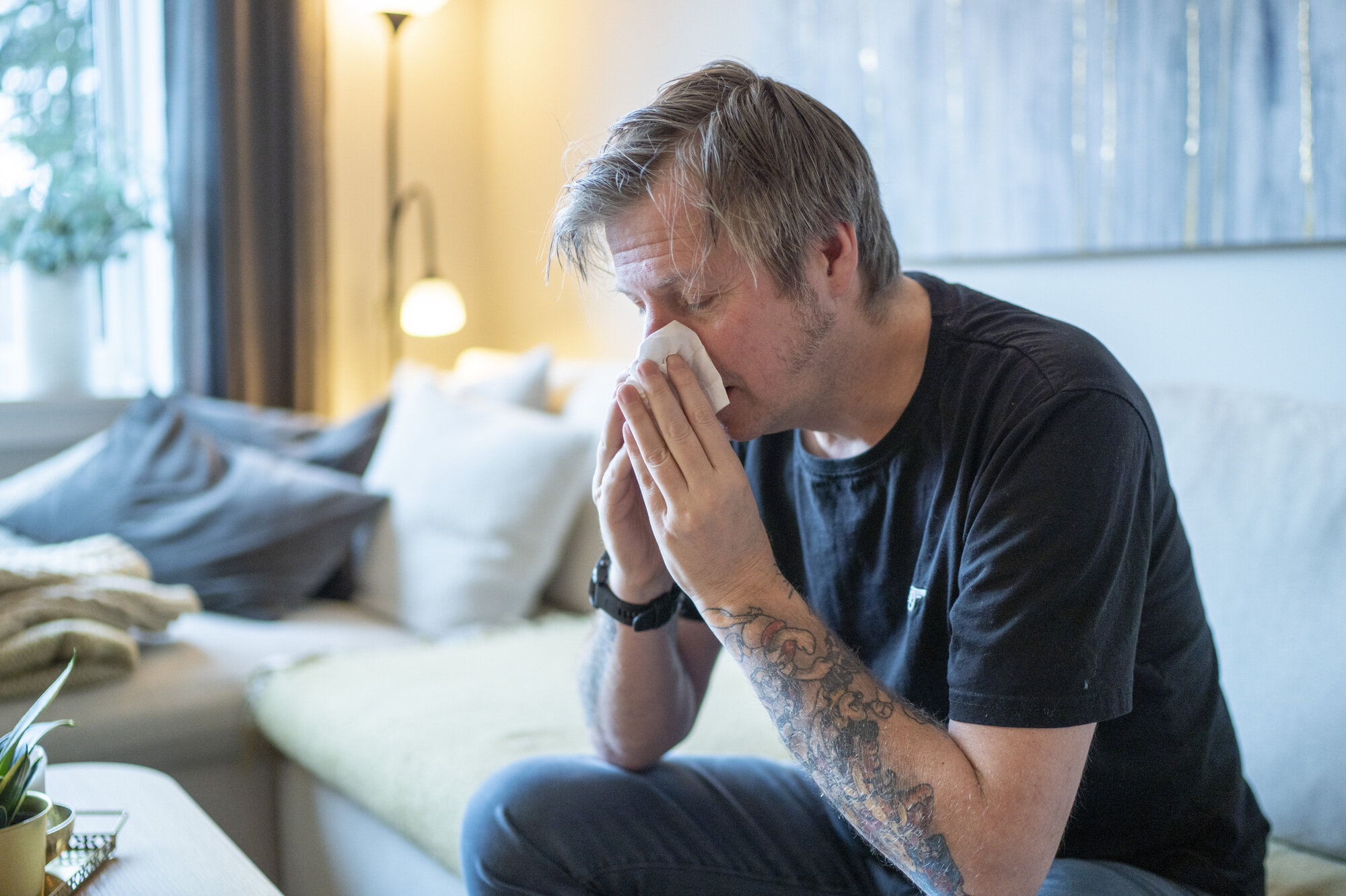 Bildet viser en mann som sitter i en sofa og snyter seg.