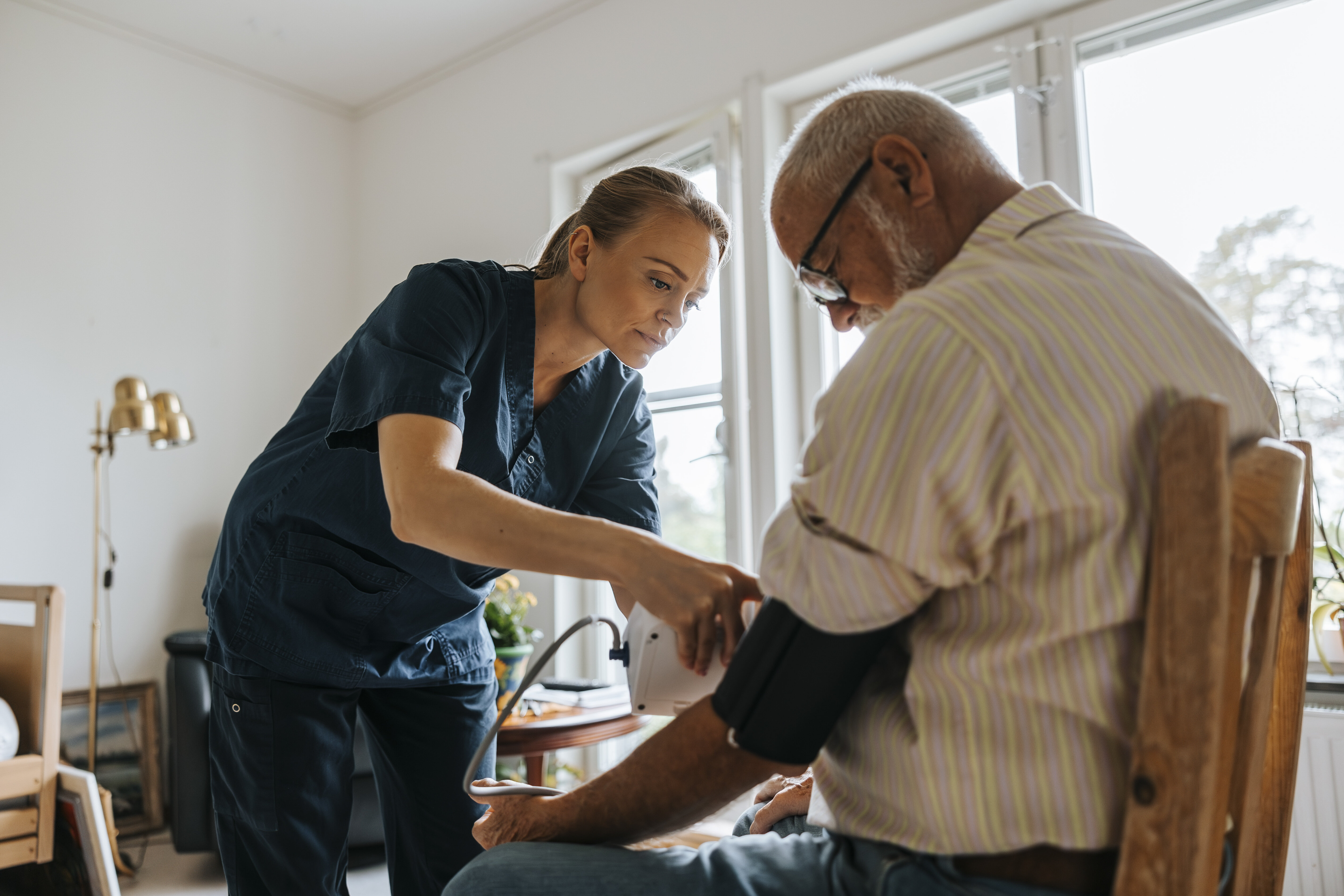 The image shows a nurse measuring the blood pressure of an elderly patient at home.