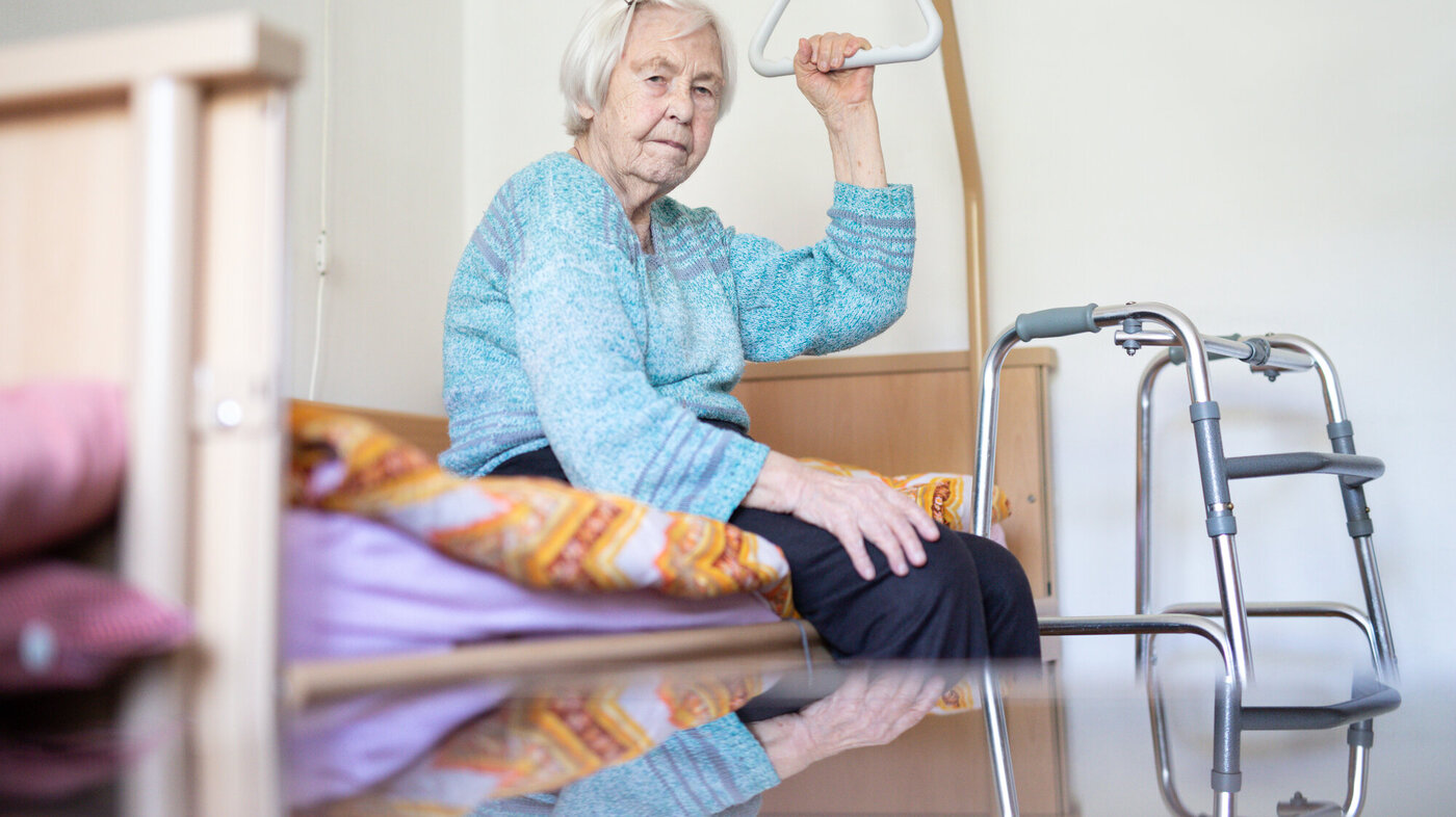 Gammel dame på pleiehjem sitter på sengen med rullator foran. Rutete pledd. 