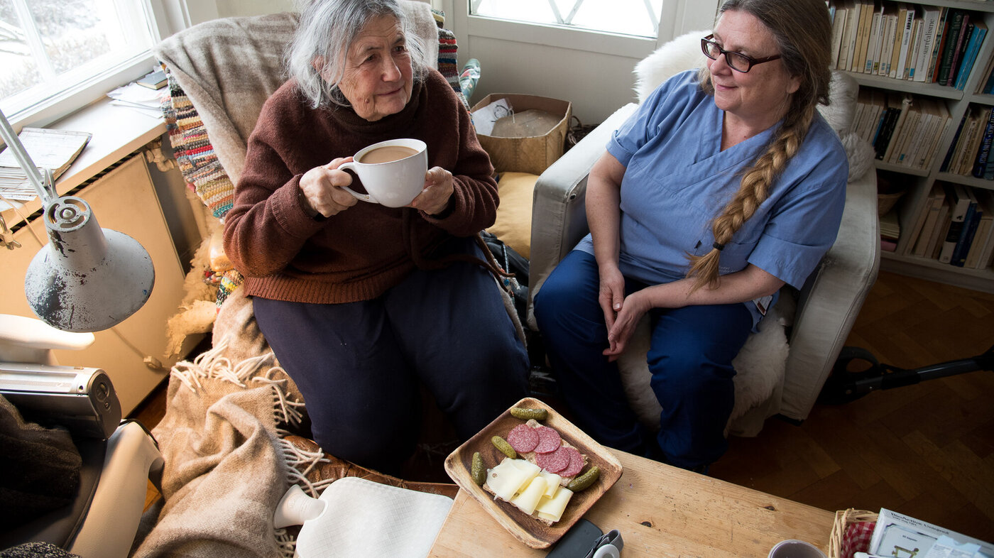 Bildet viser en beboer som drikker kaffe sammen med hjemmesykepleieren. Foran beboeren står et fat med påsmurte brødskiver.