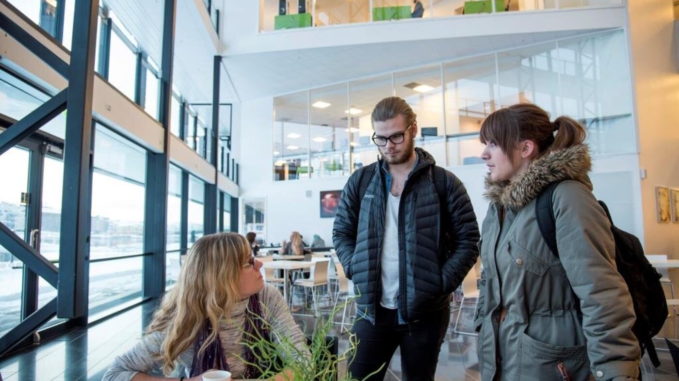 Anette Sagen, sykepleierstudent Bildet viser Anette Sagen som sitter ved et bord i kantinen og snakker med to medstudenter som står.