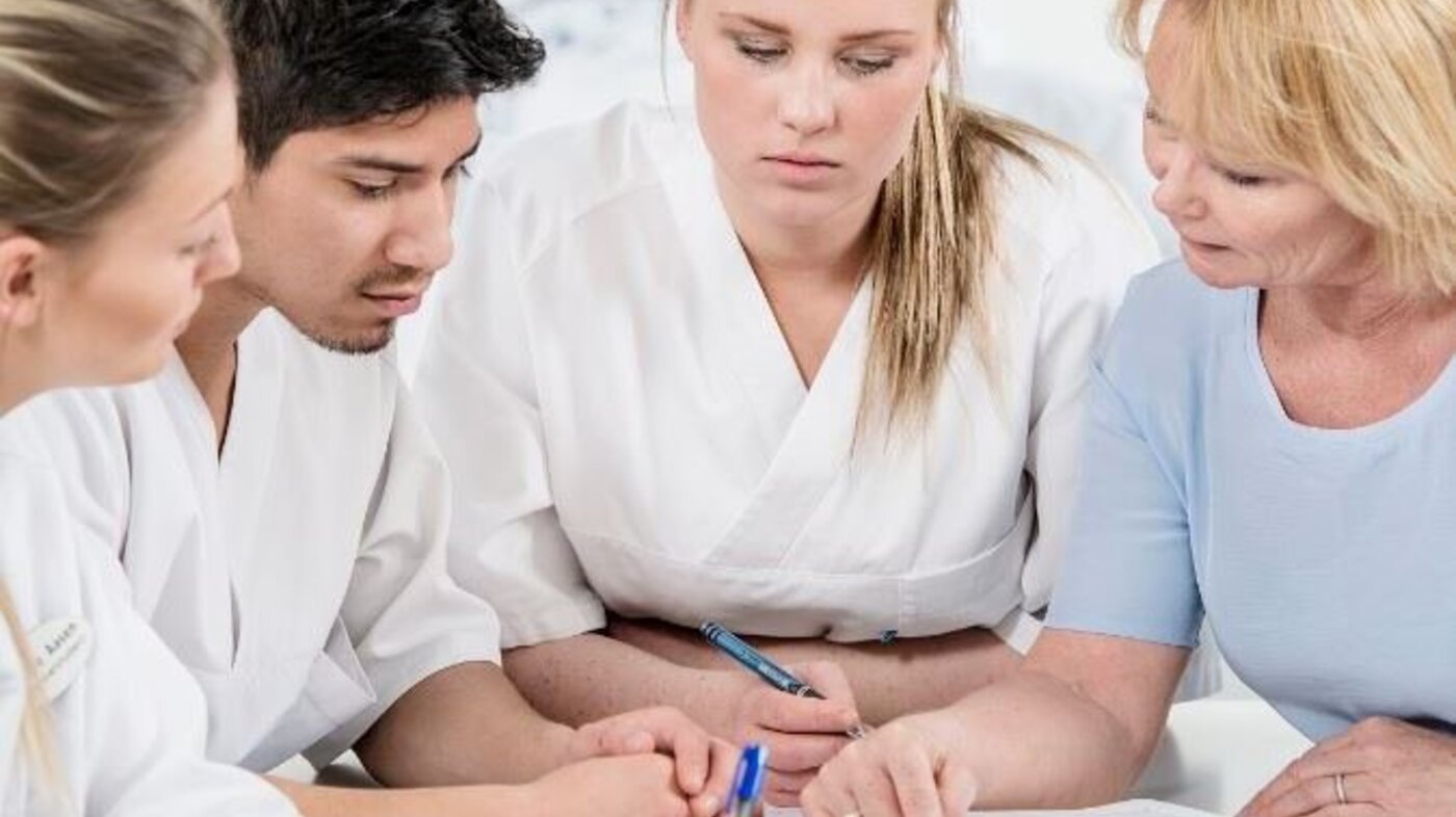 Fasilitator og studenter Fasilitator og tre studenter sitter tett sammen rundt et bord. To av dem holder i en penn, og det er papirark på bordet.