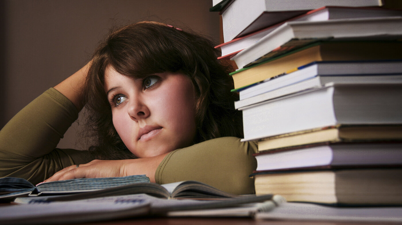 Bildet viser en student med et litt oppgitt uttrykk, som ligger over en oppslått bok på pulten. Det ligger en stabel bøker ved siden av henne.