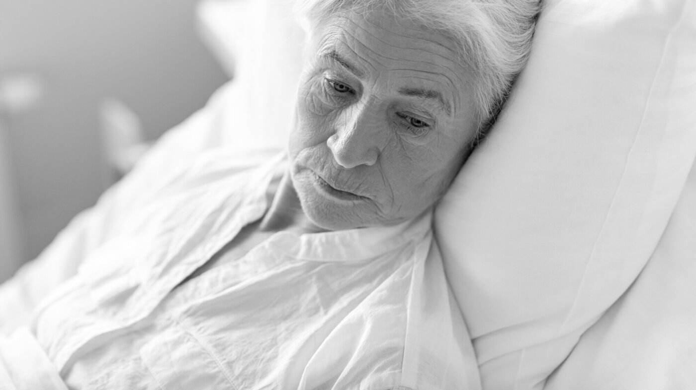 Bildet viser nærbilde av ei eldre dame i sengen, som virker ettertenksom