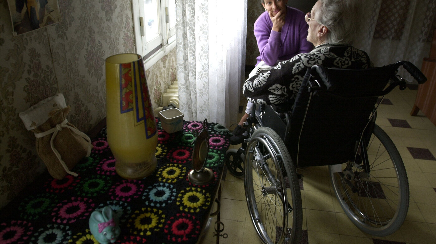 Bildet viser en eldre dame i rullestol sett bakfra og en yngre dame som sitter og lytter til henne.