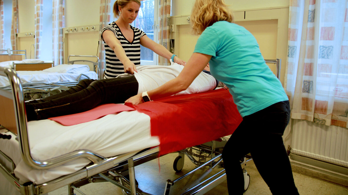 Bildet viser bedriftsfysioterapeutene Anne Aschehoug Loftu (t.v.) og Sigrunn Haug Simensen vise hvordan man kan bruke stikklaken og forflytningsduk ved forflytning av pasient i seng.