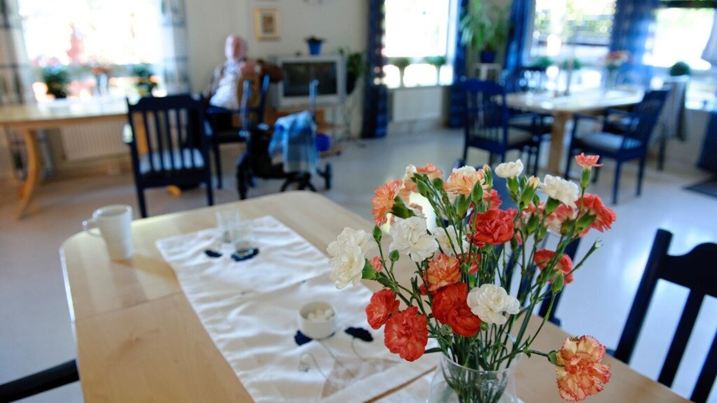 Bildet viser spisesalen på et sykehjem med blomster på bordet og en mannlig beboer i bakgrunnen